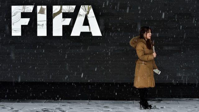A journalist stands under the snow next to a logo of the International Federation of Association Football (FIFA), at the FIFA headquarters in Zurich, during meeting of the FIFA's governing council on January 10, 2017. FIFA's ruling council on January 10, 2017 unanimously approved an expansion of the World Cup to 48 teams in 2026, with a format of 16 groups of three nations. / AFP / Michael Buholzer