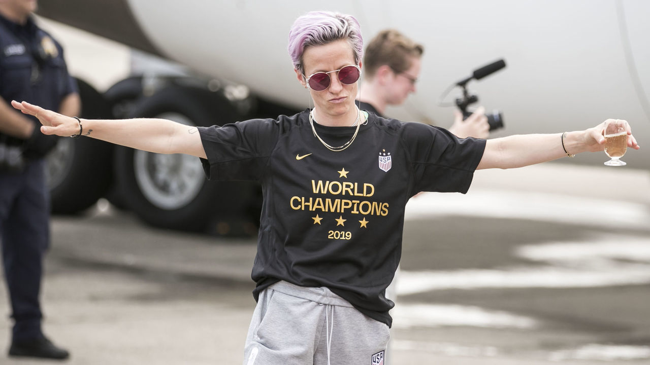 NEWARK, NJ - JULY 08: Megan Rapinoe #15 of United States with pink dyed hair holds her arms out like a plane with a drink in her hands, wearing a shirts that says World Champions as the USA Women's National team arrives back in the U.S. after winning their 4th FIFA World Cup title against Netherlands at Newark Liberty International Airport on July 08, 2019 in Newark, NJ, USA.