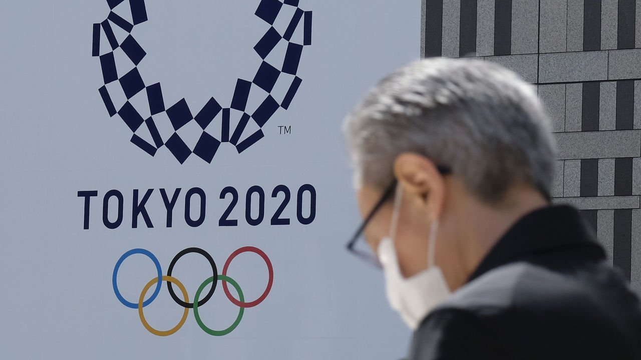 A man wearing a face mask walks before the logo of the Tokyo 2020 Olympic Games displayed on the Tokyo Metropolitan Government building in Tokyo on March 24, 2020. - The International Olympic Committee came under pressure to speed up its decision about postponing the Tokyo Games on March 24 as athletes criticised the four-week deadline and the United States joined calls to delay the competition.