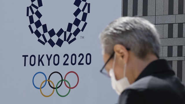 A man wearing a face mask walks before the logo of the Tokyo 2020 Olympic Games displayed on the Tokyo Metropolitan Government building in Tokyo on March 24, 2020. - The International Olympic Committee came under pressure to speed up its decision about postponing the Tokyo Games on March 24 as athletes criticised the four-week deadline and the United States joined calls to delay the competition.