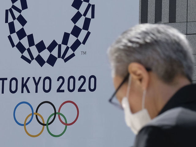 A man wearing a face mask walks before the logo of the Tokyo 2020 Olympic Games displayed on the Tokyo Metropolitan Government building in Tokyo on March 24, 2020. - The International Olympic Committee came under pressure to speed up its decision about postponing the Tokyo Games on March 24 as athletes criticised the four-week deadline and the United States joined calls to delay the competition.