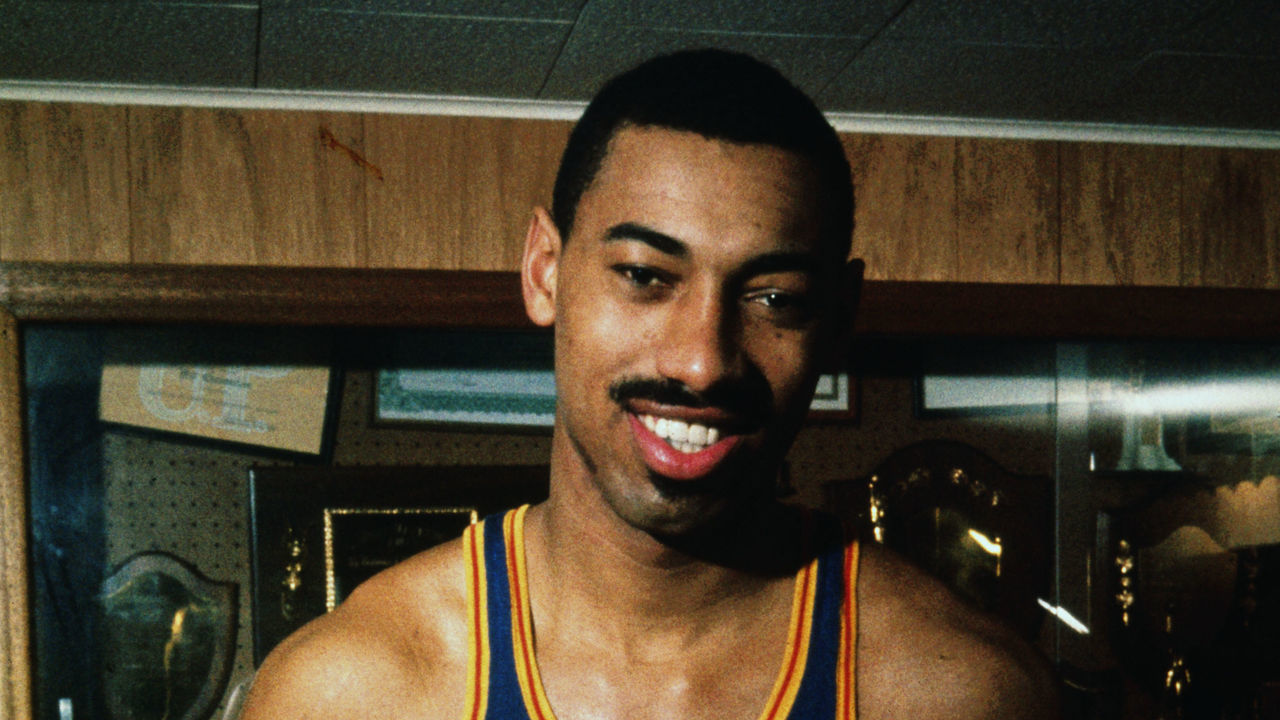 Philadelphia Warriors basketball player Wilt Chamberlain holds a basketball while leaning against a trophy case.