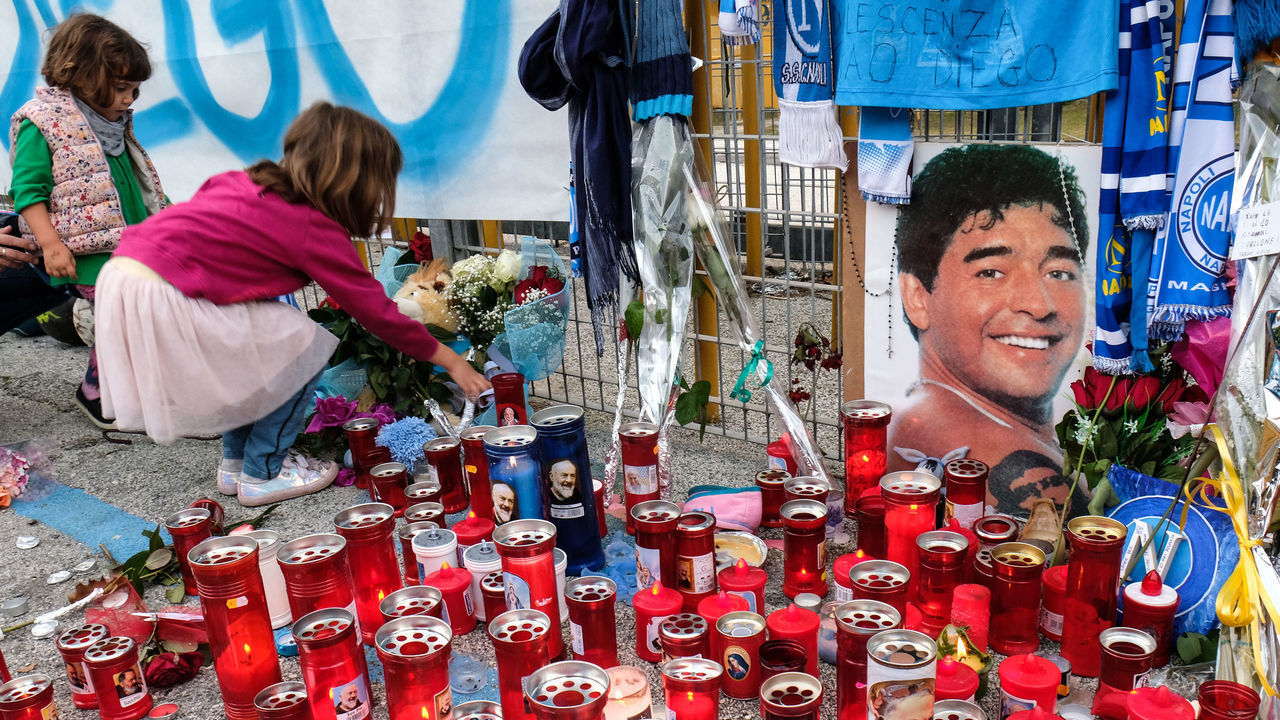 NAPLES, CAMPANIA, ITALY - 2020/11/28: A little girl places a flower near candle and memorabilia give in honour by supporters of the football legend Diego Armando Maradona are placed outside the San Paolo Stadium.