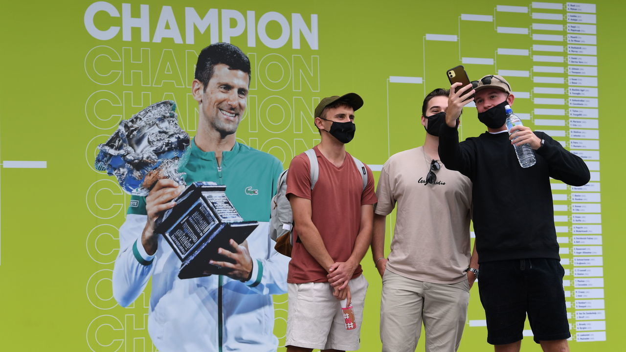 MELBOURNE, AUSTRALIA - JANUARY 17: Spectators take selfies as they arrive at Melbourne Park with an image of 2021 champion Novak Djokovic of Serbia during day one of the 2022 Australian Open at Melbourne Park on January 17, 2022 in Melbourne, Australia.
