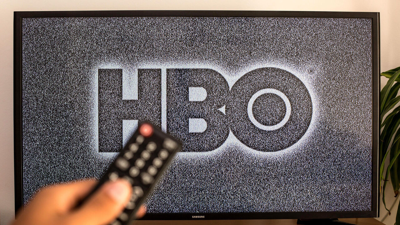 SPAIN - 2021/07/13: In this photo illustration a close-up of a hand holding a TV remote control seen displayed in front of the HBO logo on a tv in the background. (Photo Illustration by Thiago Prudencio/SOPA Images/LightRocket via Getty Images)