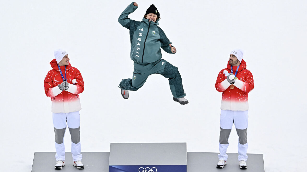 Brazil's gold medalist Lucas Pinheiro Braathen (C) leaps onto the podium flanked by Switzerland's silver medalist Marco Odermatt (L), and Switzerland's bronze medalist Loic Meillard during the podium of the men's giant slalom alpine skiing event during the Milano Cortina 2026 Winter Olympic Games at the Stelvio Ski Centre in Bormio (Valtellina) on February 14, 2026.
