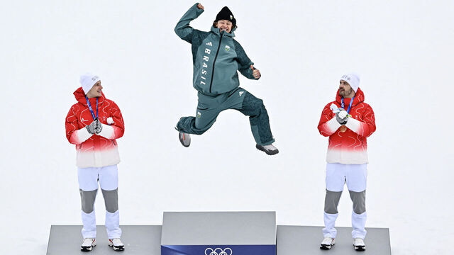 Brazil's gold medalist Lucas Pinheiro Braathen (C) leaps onto the podium flanked by Switzerland's silver medalist Marco Odermatt (L), and Switzerland's bronze medalist Loic Meillard during the podium of the men's giant slalom alpine skiing event during the Milano Cortina 2026 Winter Olympic Games at the Stelvio Ski Centre in Bormio (Valtellina) on February 14, 2026.